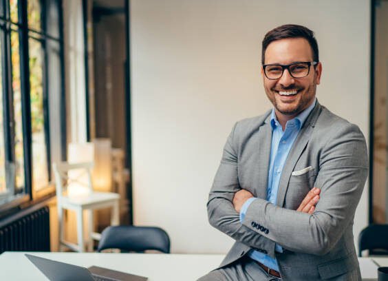 Portrait of young happy businessman wearing grey suit and blue shirt standing in his office and smiling with arms crossed